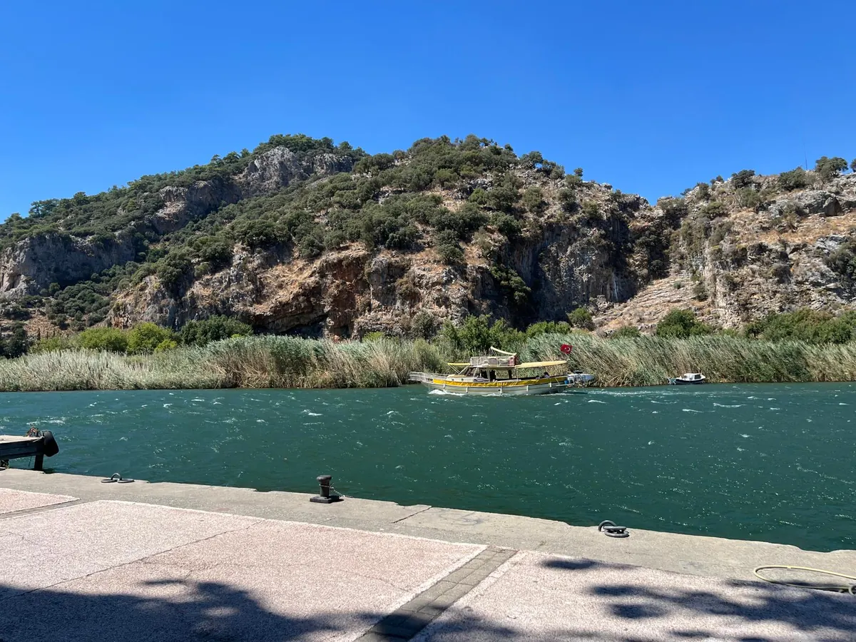 A Dalyan tour boat on the turquoise river beside tall reed beds, below the pine-covered limestone cliffs of the delta