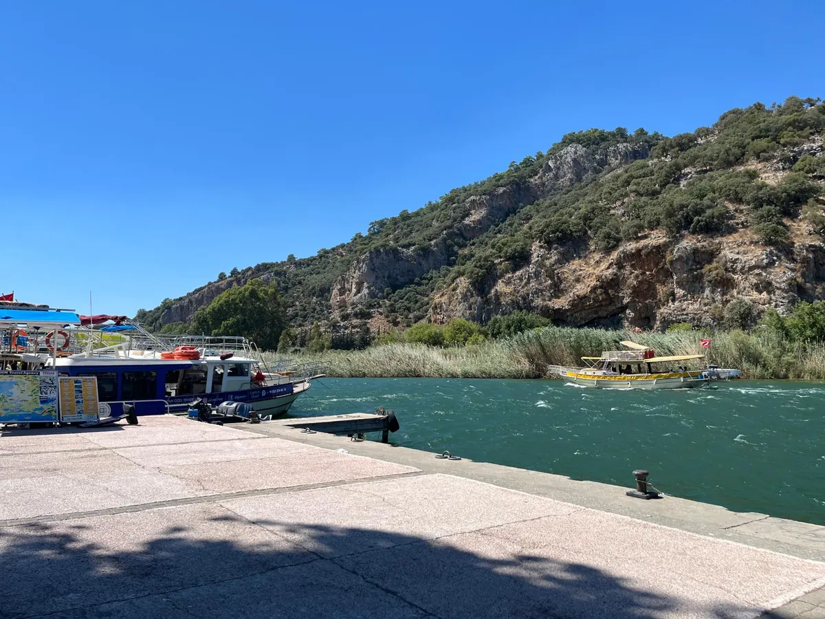 Tour boats moored along the Dalyan town quay with the day’s trip boards on the jetty and a boat heading up the river