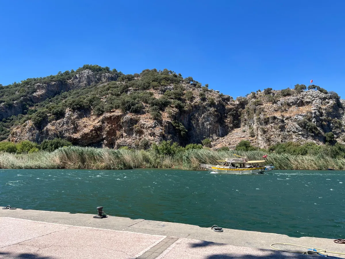 A traditional yellow-awning Dalyan tour boat passing the reed beds on the Dalyan river, with the limestone cliffs of Kaunos rising behind