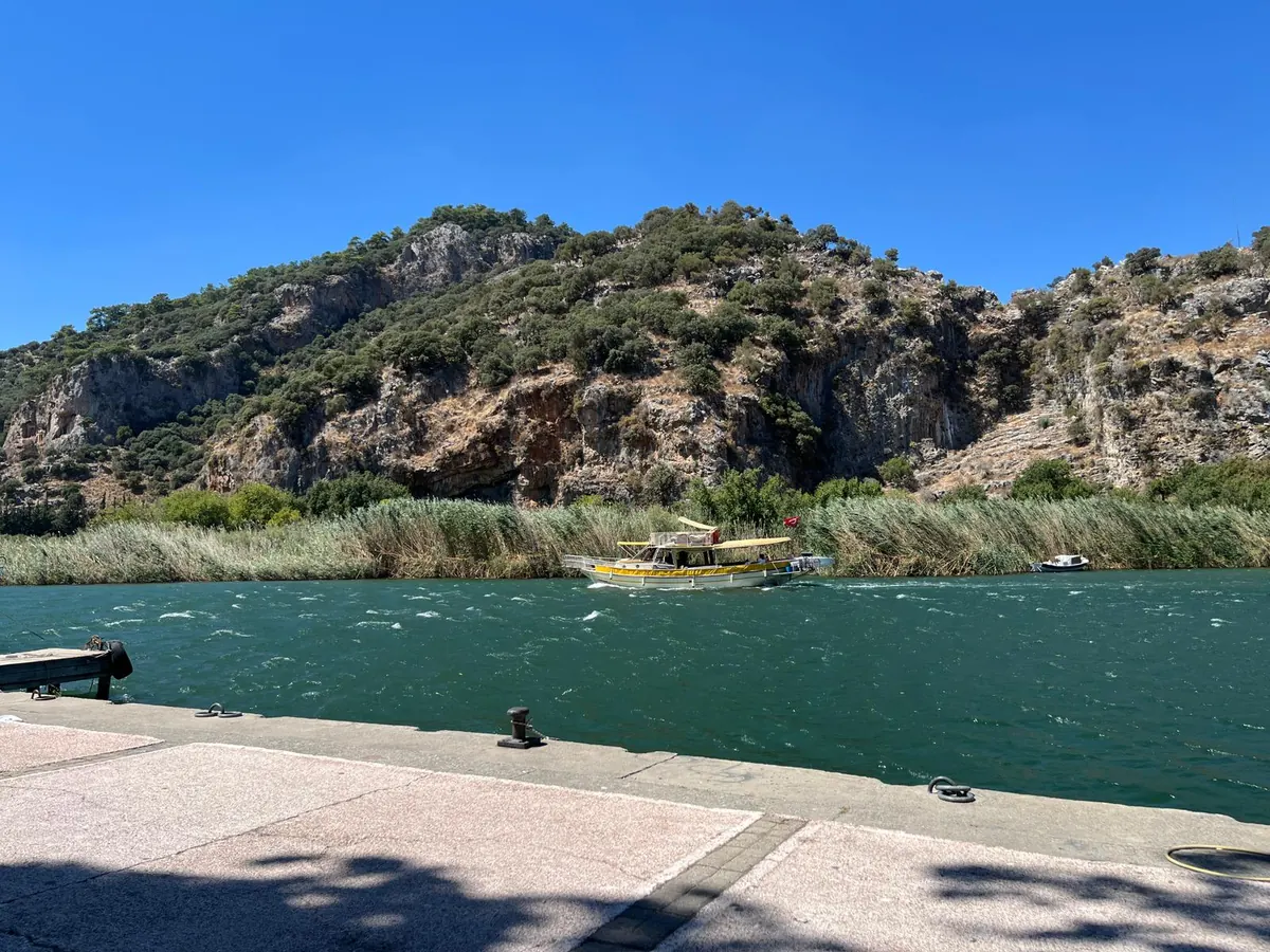 The limestone cliff of ancient Kaunos above the Dalyan river, with the rock-cut tombs on the left face and a tour boat passing the reeds
