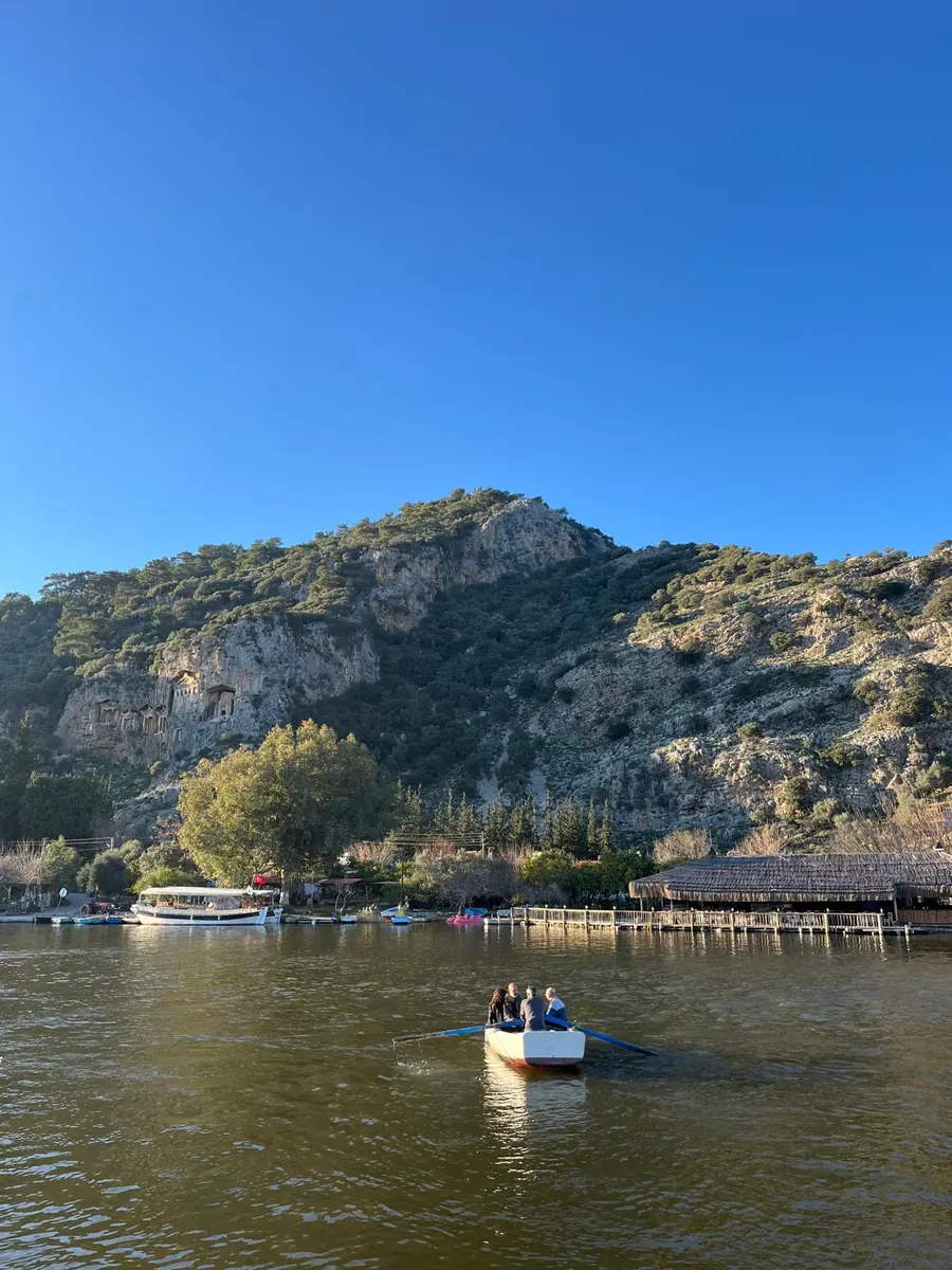 A small rowing boat crossing the Dalyan river below the Kaunos rock tombs, with the jetties and riverside restaurants behind